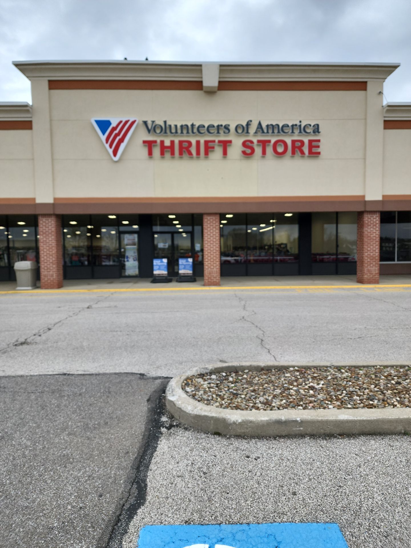 0118-Apr 10 2024 10_55am-fiyB Exterior view of a Volunteers of America Thrift Store with glass doors and windows, located in a shopping plaza on a cloudy day.
