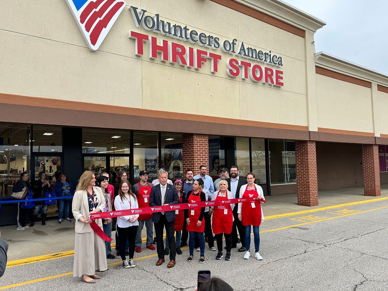 32-Apr-18-2024-07_36am-72Mz A group of people stands in front of a Volunteers of America Thrift Store, holding a red ribbon for a ribbon-cutting ceremony.