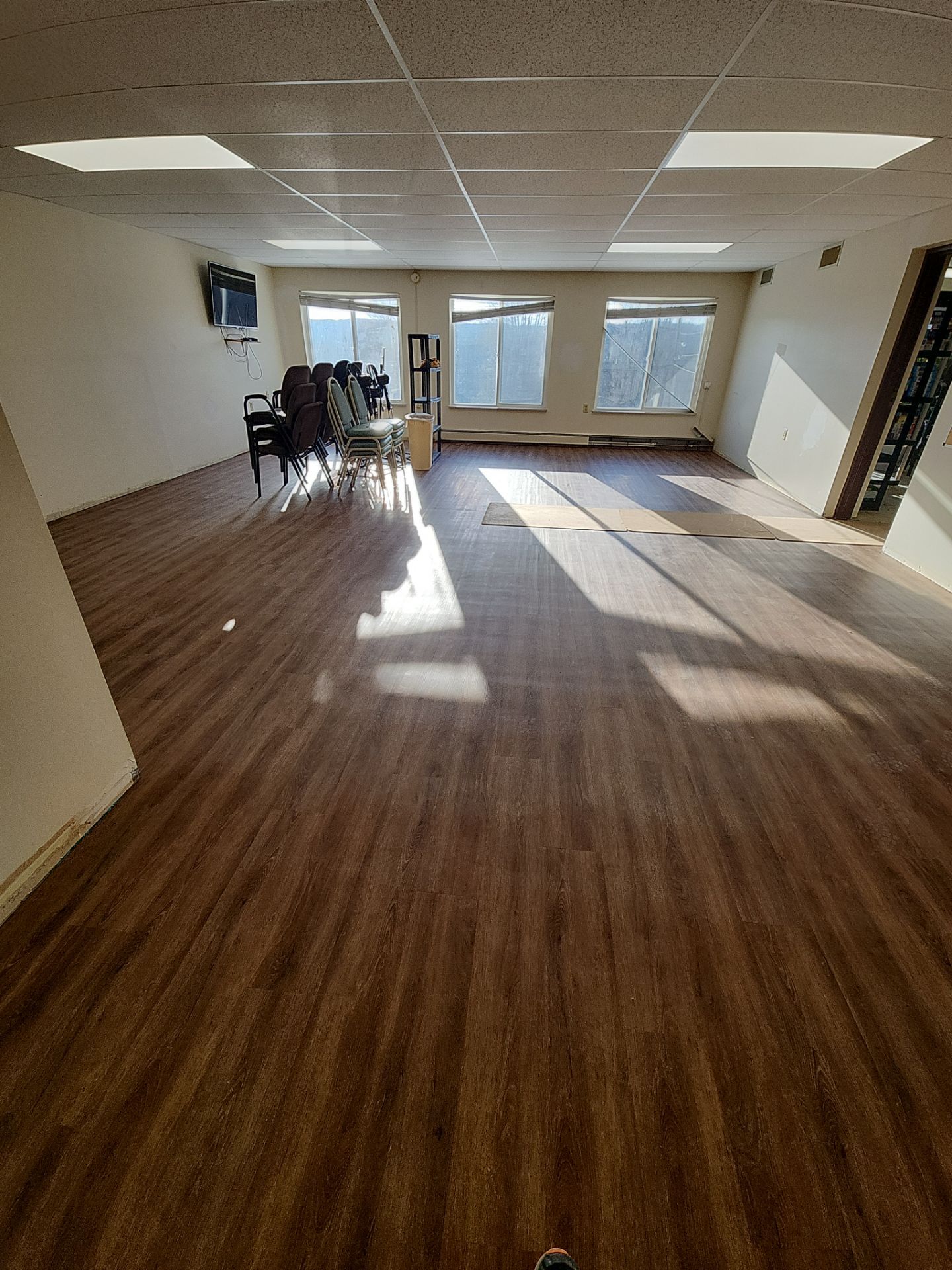A mostly empty room with wood flooring, stacked chairs, a TV mounted on the wall, and large windows letting in sunlight.