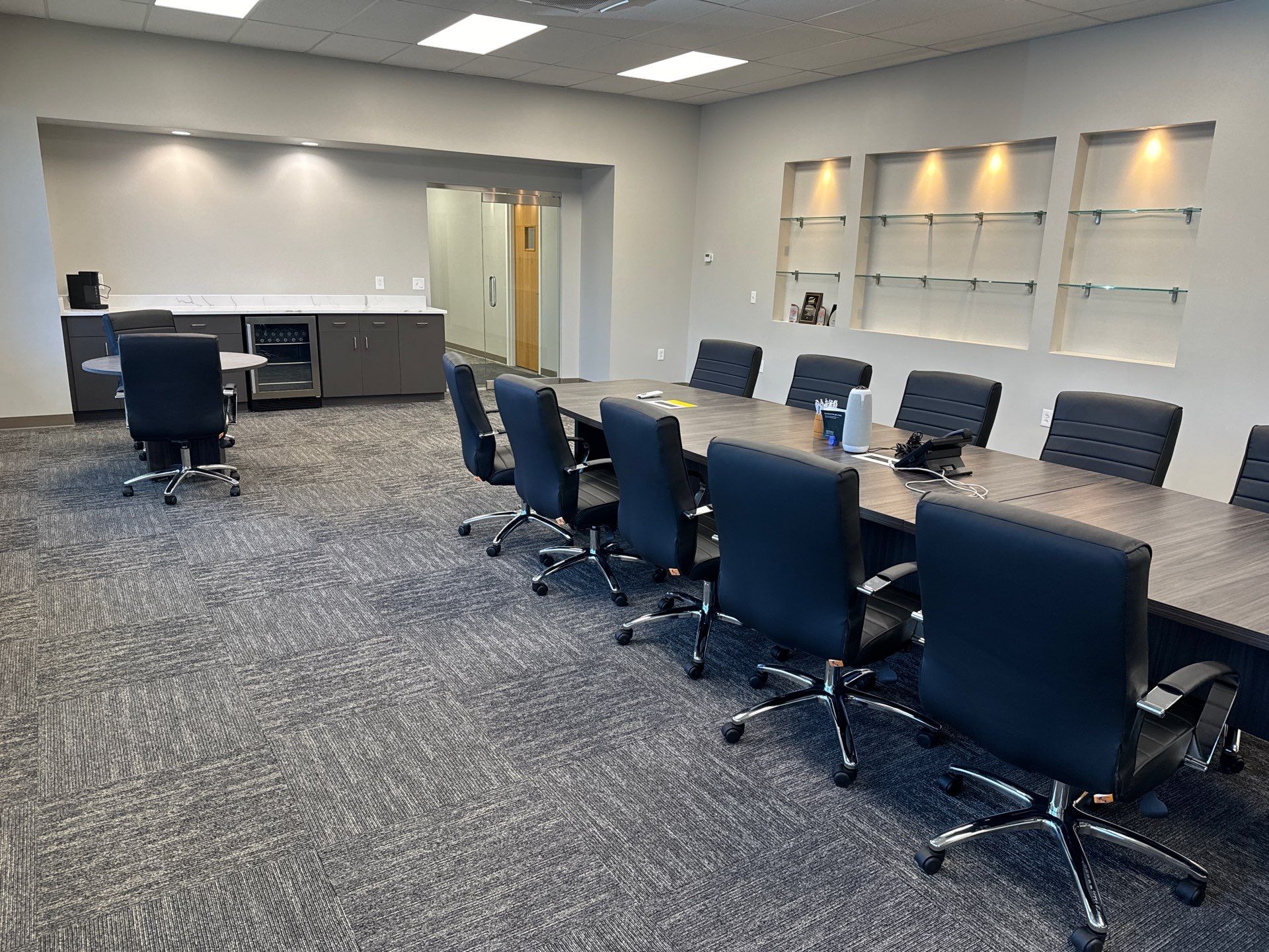A modern conference room with a long table, black chairs on wheels, built-in shelves with spotlights, and a side counter with a coffee maker.