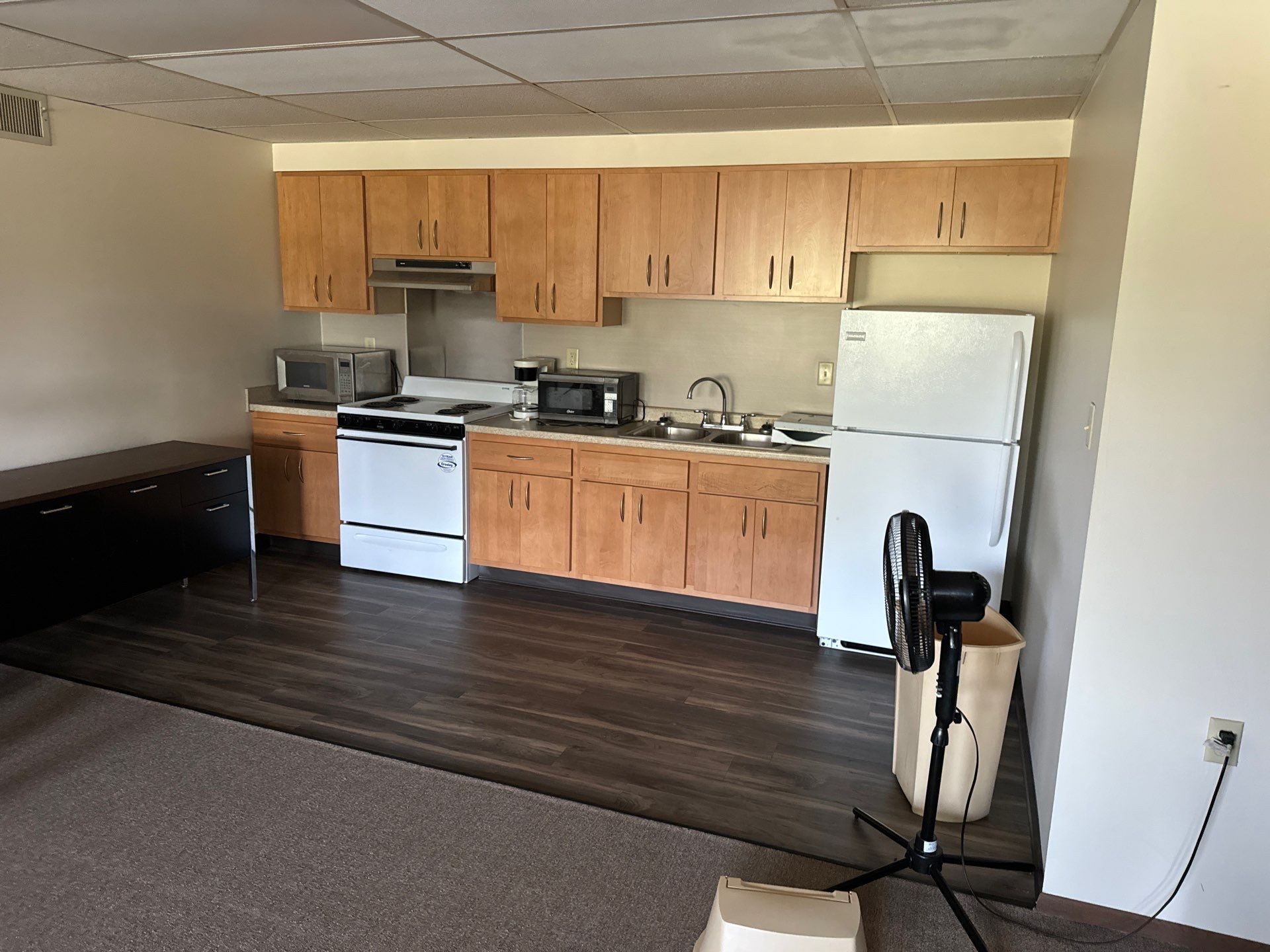 9-Sep 13, 2024 14-16-35-go99 A small kitchen with wooden cabinets, white appliances including a stove and refrigerator, two microwaves, a sink, and a black floor fan in the foreground.