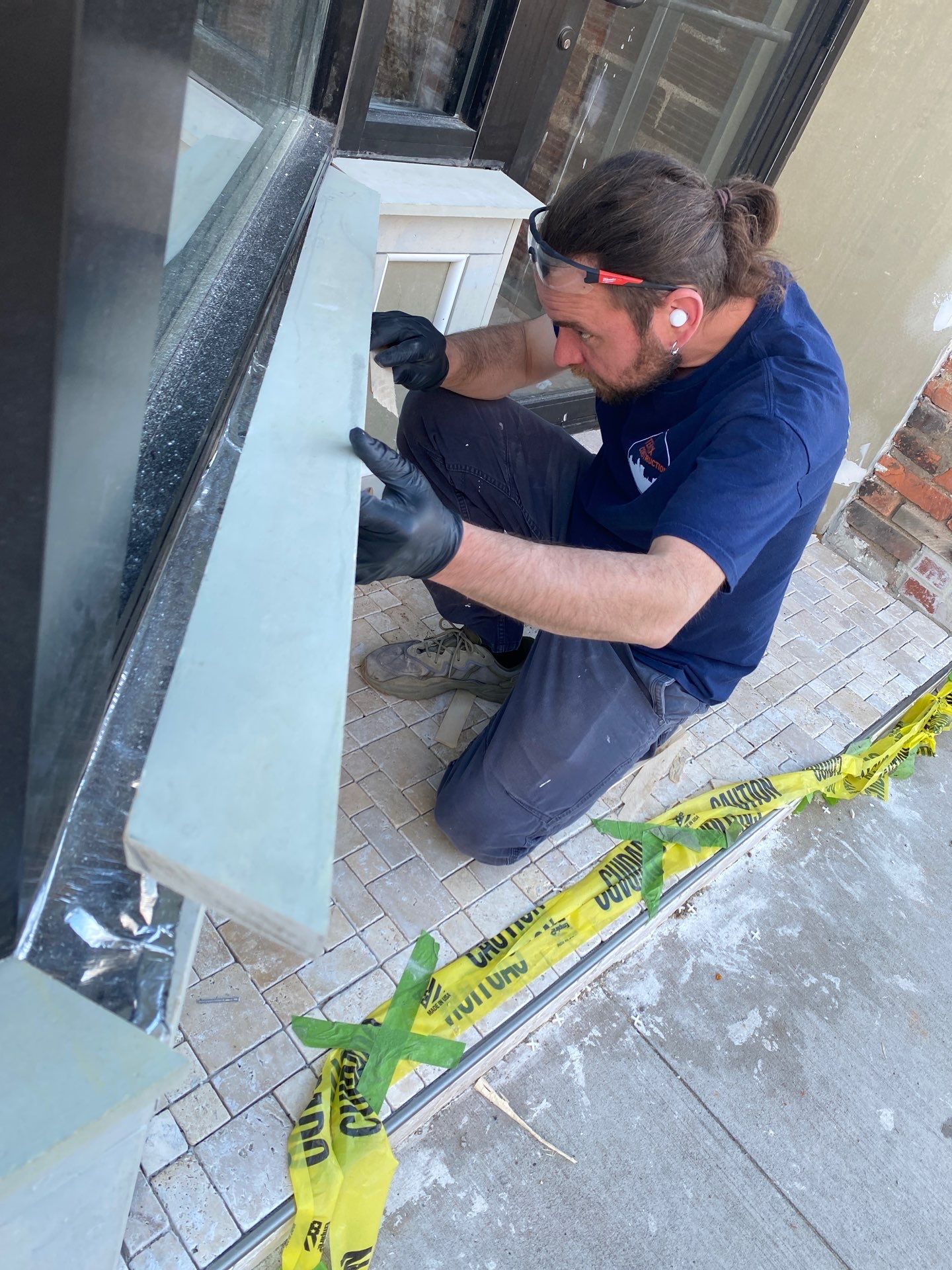 CompanyCam 2227319024 A man wearing gloves and ear protection works on a window frame outside a building, kneeling on a tiled surface behind yellow caution tape.