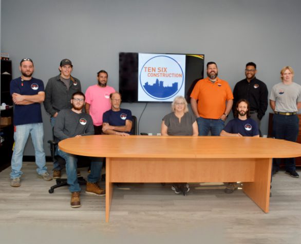 Ten people pose for a group photo in an office, seated and standing around a wooden table with a Ten Six Construction logo displayed on a screen behind them.
