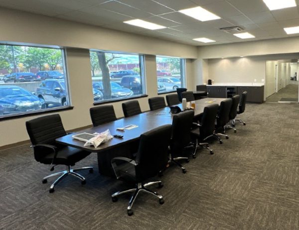 A modern conference room with a long table, ten black chairs, large windows showing a parking lot, and a counter with a coffee maker in the back.