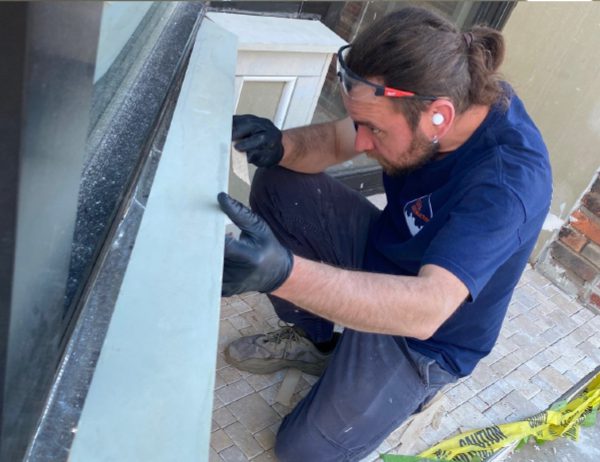 A man wearing gloves and earplugs installs a panel on an exterior window while kneeling on a tiled surface.