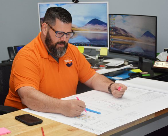 A man wearing an orange shirt and glasses marks architectural plans with colored pens at a desk with computer monitors and office supplies.