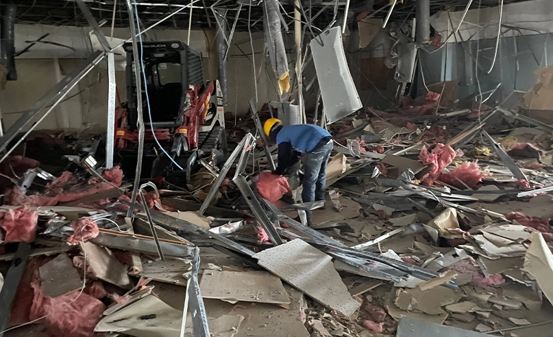 A worker wearing a yellow helmet clears debris in a damaged building interior with exposed insulation, metal, and drywall, alongside a small construction vehicle.
