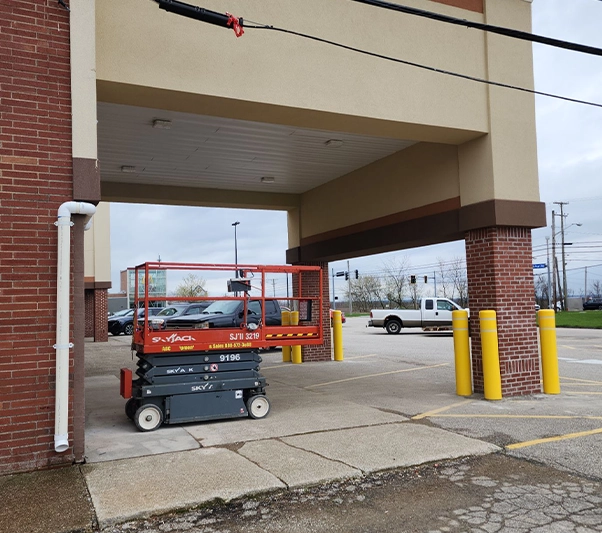 A red and gray scissor lift is parked under a covered area outside a building near yellow bollards and a parking lot.