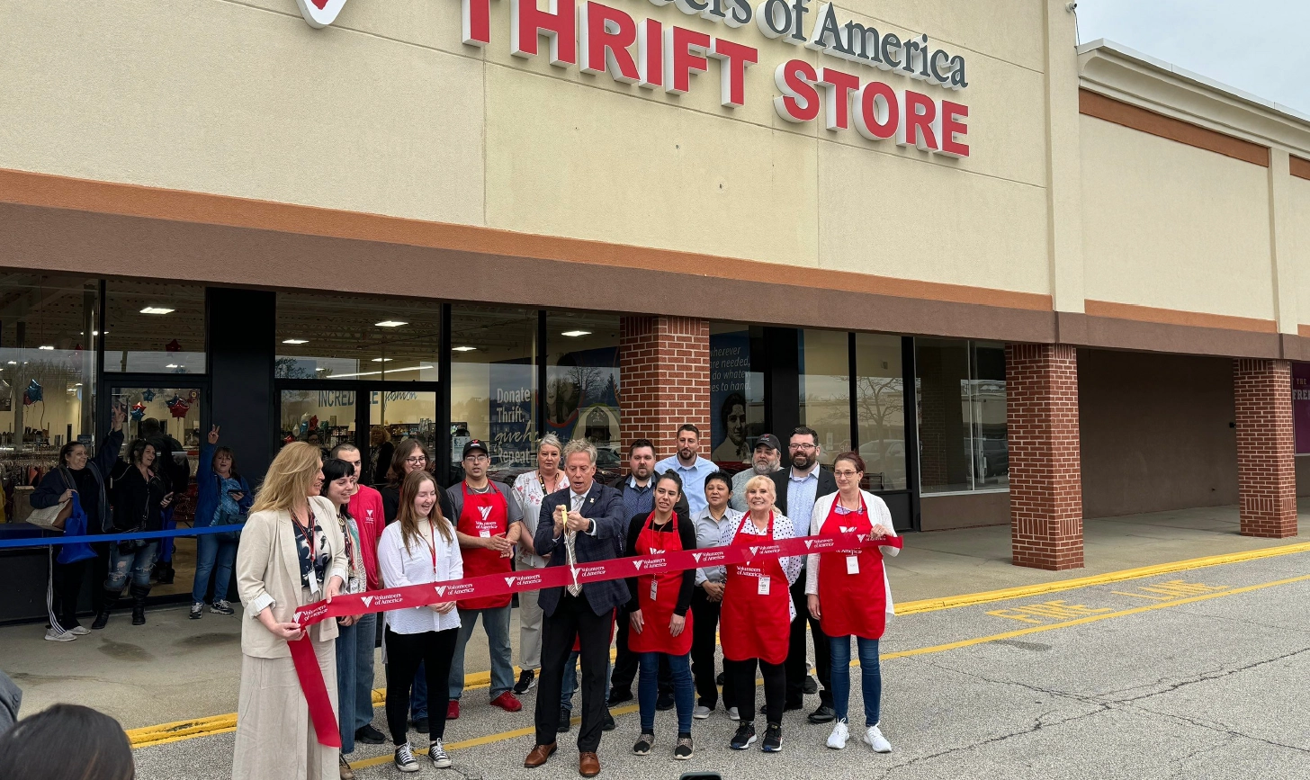 A group of people, some wearing red aprons, participate in a ribbon-cutting ceremony outside a thrift store with a "Thrift Store" sign above the entrance.