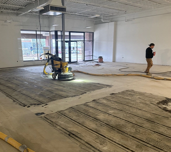 A worker operates a floor grinding machine in a large, empty commercial space with exposed ceilings and unfinished floors.