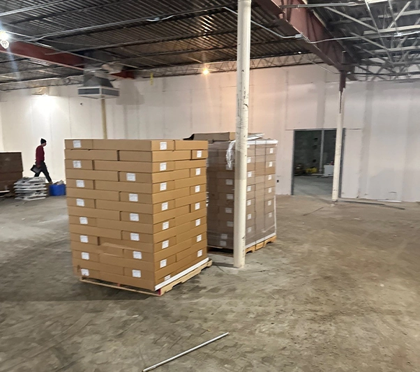 Two large pallets stacked with cardboard boxes in a spacious, mostly empty warehouse with a person walking in the background.