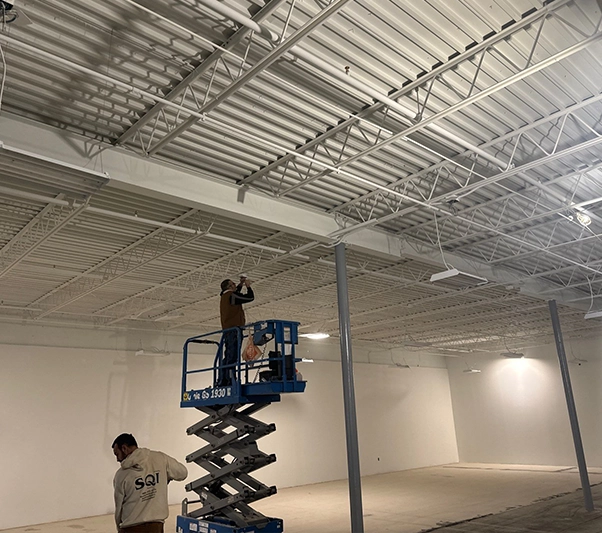 Two workers are in a large, mostly empty industrial space; one is on a scissor lift working on the ceiling, while the other stands on the ground.