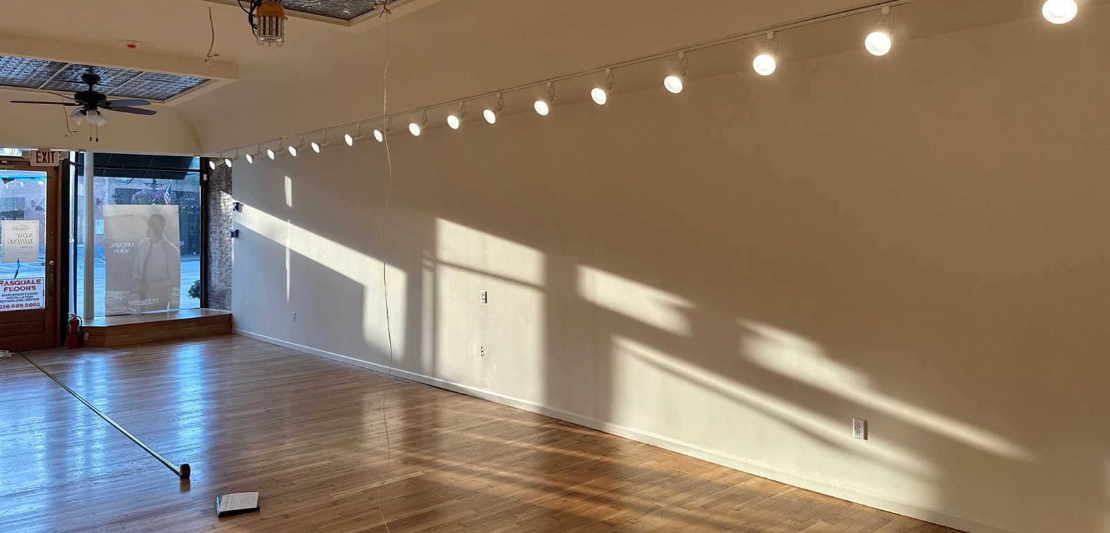 Empty room with wooden floor, cream-colored walls, track lighting on the ceiling, and sunlight casting shadows through large front windows near the entrance.