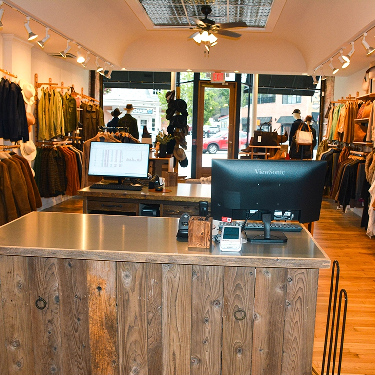 A wooden checkout counter with two monitors inside a clothing store, with racks of clothes and hats on either side and large windows at the front.
