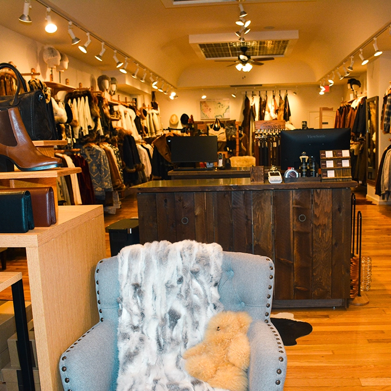 Clothing boutique interior with wooden floors, racks of clothes, shelves with accessories, a cash register counter, and a cushioned chair draped with a fur throw and a teddy bear.