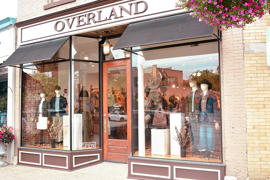 Storefront of Overland with large glass windows displaying mannequins dressed in casual clothing and leather accessories; a hanging flower basket is visible on the right.