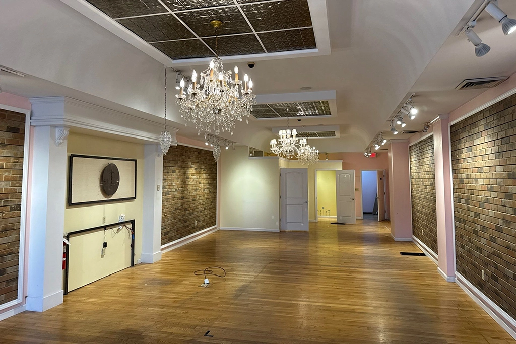 Empty room with wood floors, exposed brick walls, chandeliers, and recessed lighting; some wires and wall mounts are visible on the left wall.