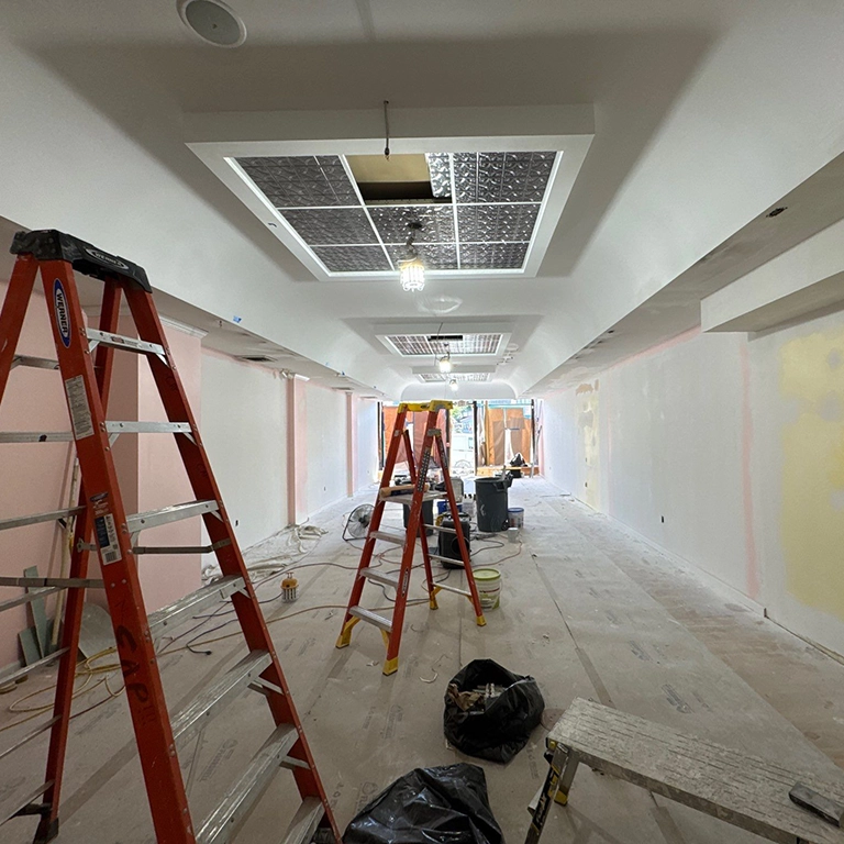 Room under renovation with ladders, exposed ceiling panels, construction materials, and unfinished walls. Natural light is visible from windows at the far end.