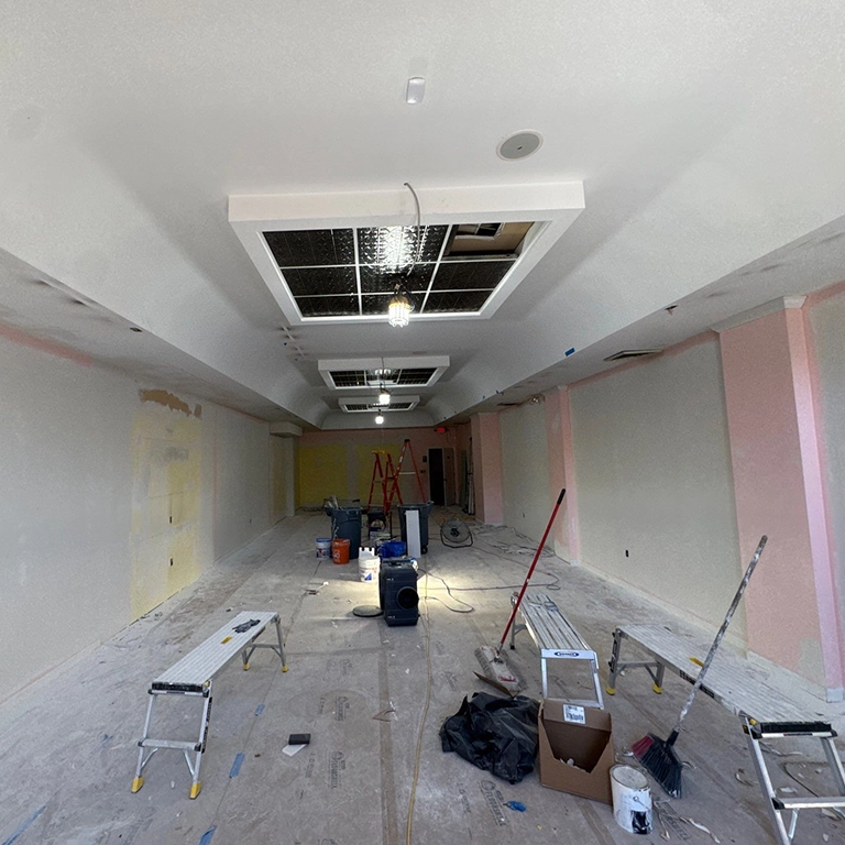 Long room under renovation with construction tools, ladders, and benches scattered across the floor; ceiling panels are open with visible wiring and unfinished walls.
