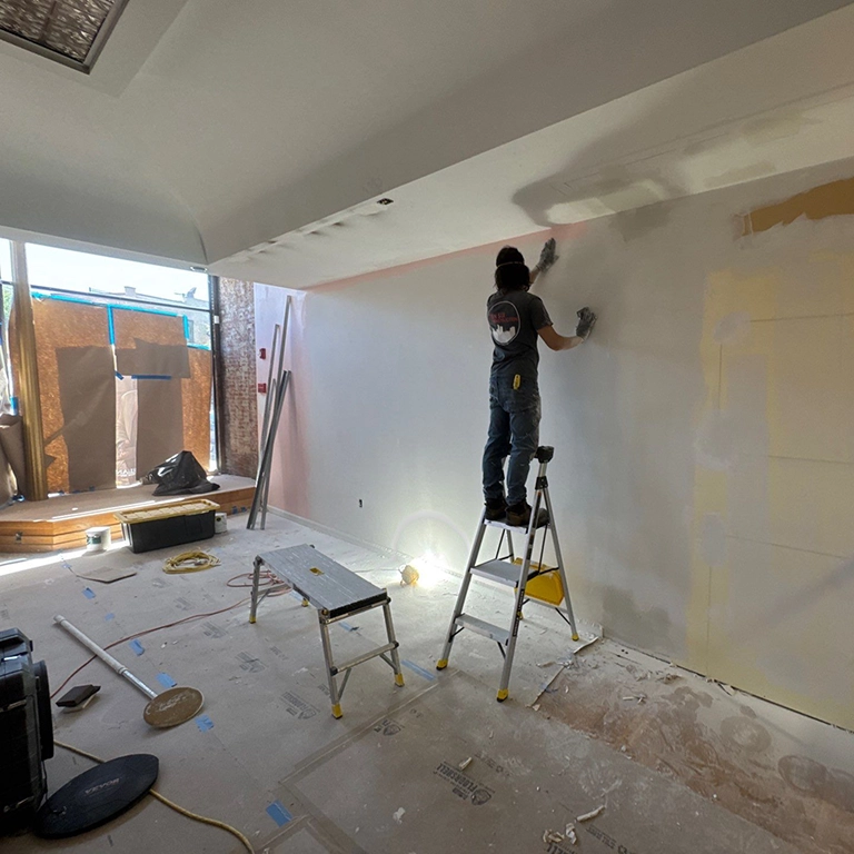A person stands on a ladder applying plaster or paint to a large wall in a partially renovated room with construction materials scattered on the floor.