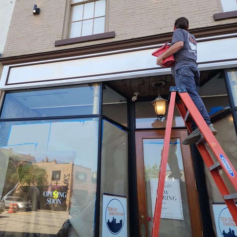 A person stands on a red ladder painting a white sign above the entrance of a storefront with "Now Hiring" and "Opening Soon" signs in the windows.