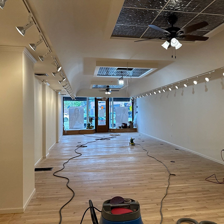 An empty, newly renovated room with light wood floors, ceiling fans, track lighting, and cleaning equipment scattered across the floor. Large front windows let in natural light.