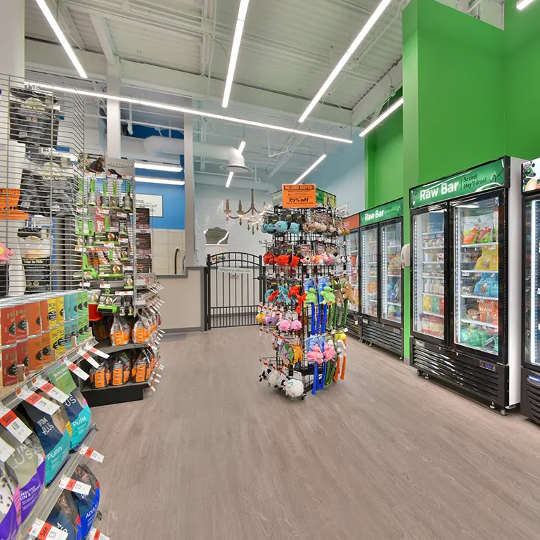 Pet store interior with shelves of pet food, toys, and supplies, plus refrigerated sections on the right and a fenced area in the back. Bright lighting and green accent walls.