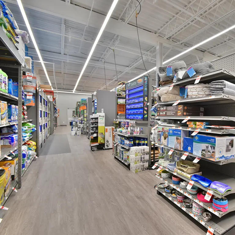Aisle in a pet supply store with shelves stocked with pet food, beds, feeding bowls, and accessories under bright overhead lighting.