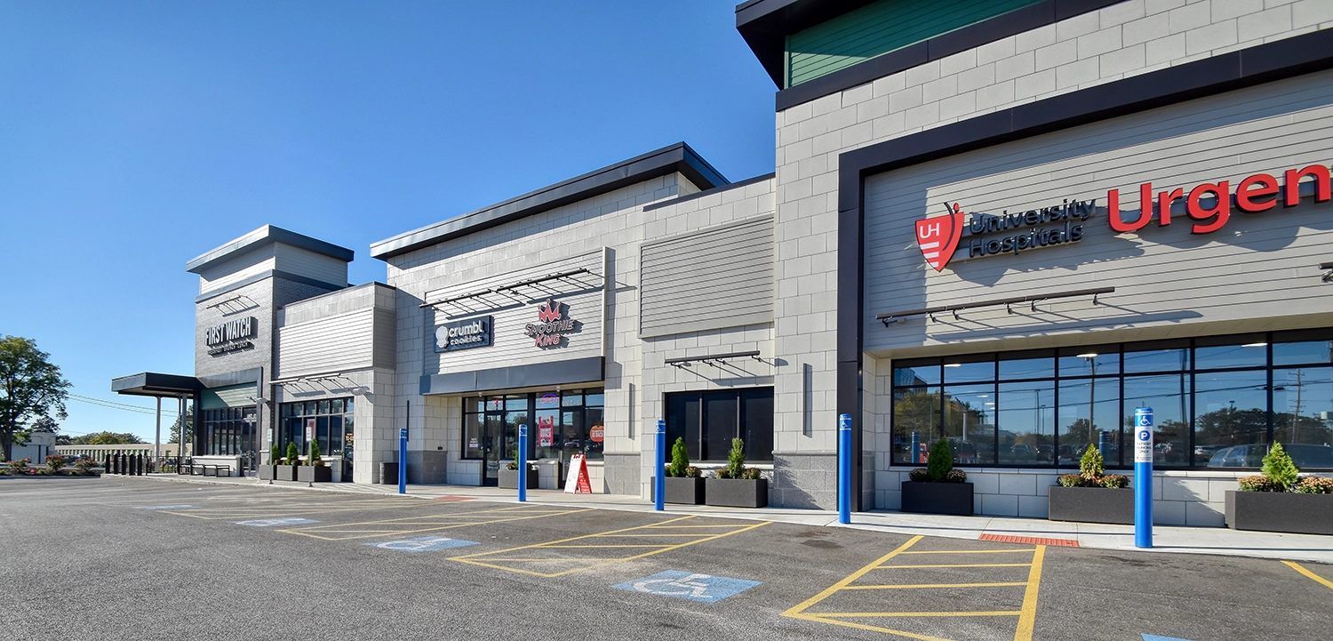 A modern shopping plaza with several storefronts, including a medical urgent care, visible on a sunny day. Parking spaces, including accessible spots, are in front.
