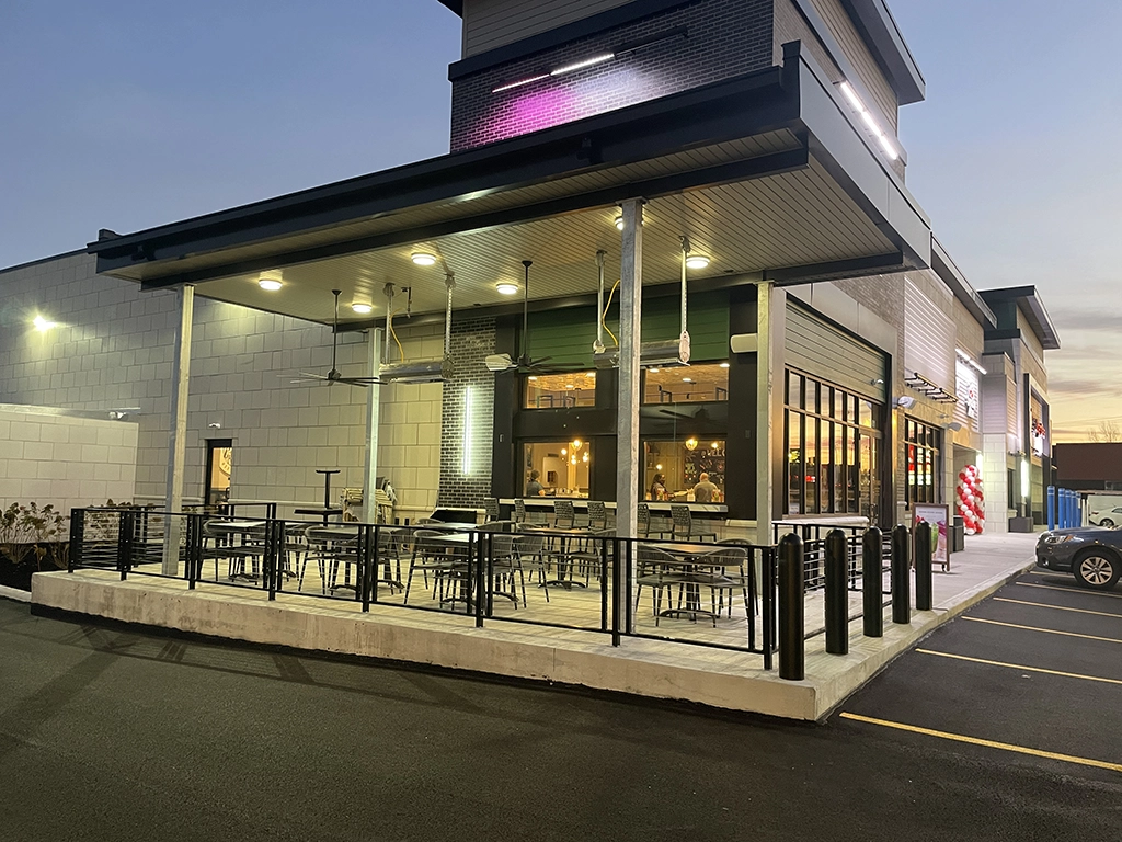 Outdoor patio with empty tables and chairs outside a modern restaurant at dusk, illuminated by overhead lights, with parked cars visible on the side.