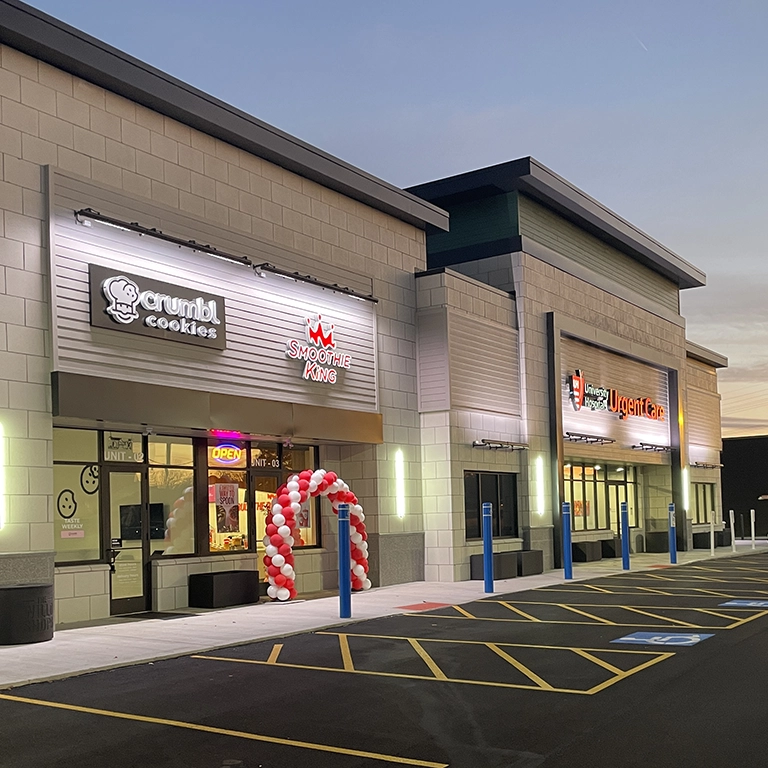 A modern shopping plaza at sunset with Crumbl Cookies, Smoothie King, and Pizza Yogurt Café storefronts; a red and white balloon arch is at one entrance.