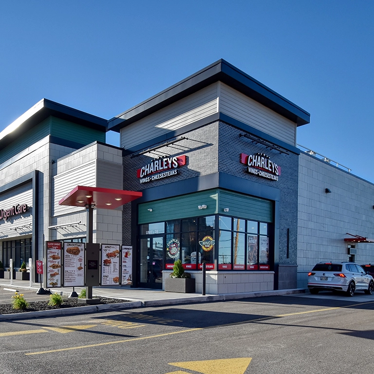 A Charleys cheesesteaks and wings restaurant with a drive-thru lane, menu boards, and a car parked near the pickup window on a clear day.