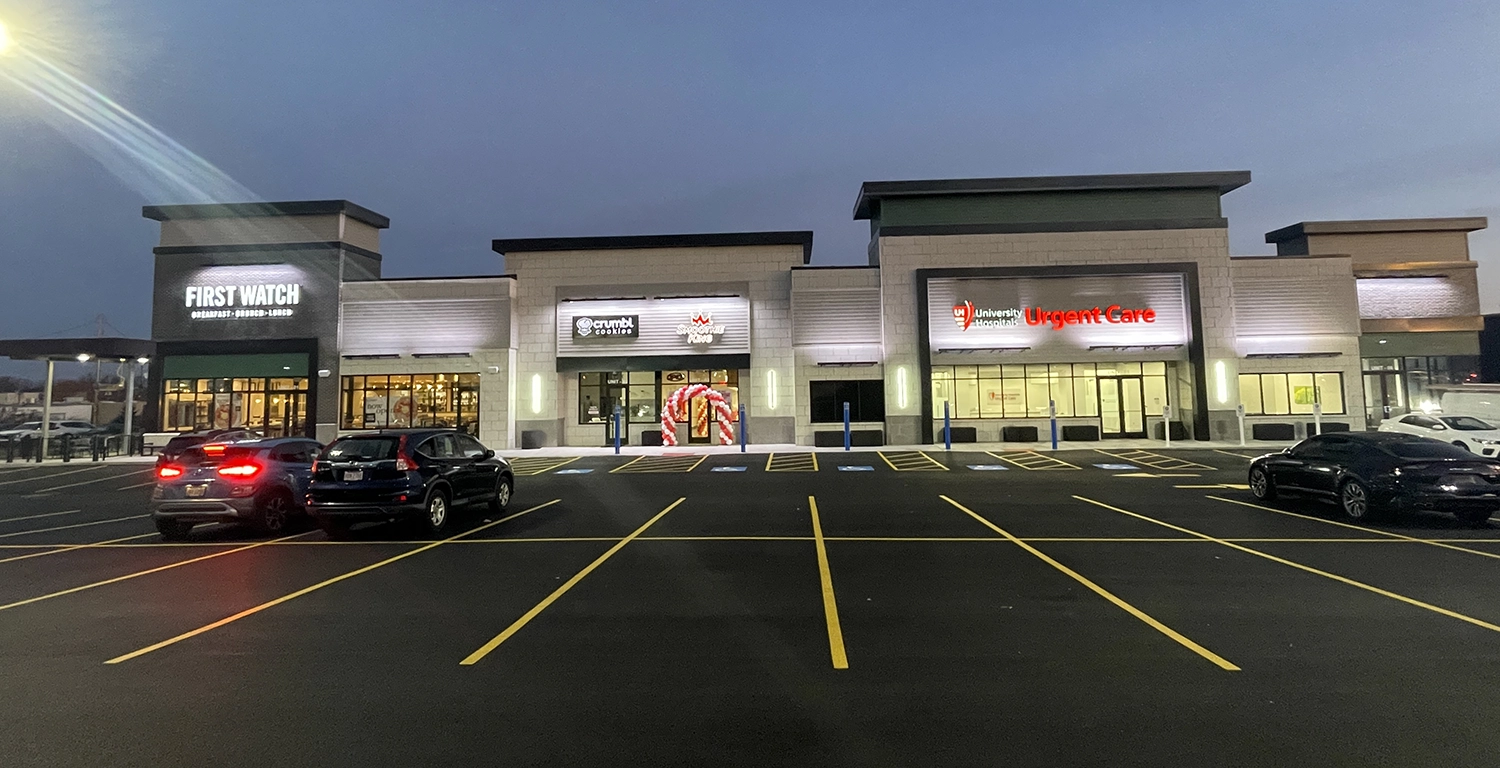 A strip mall at dusk with First Watch, a dental office, and an urgent care center visible; several cars are parked in front.