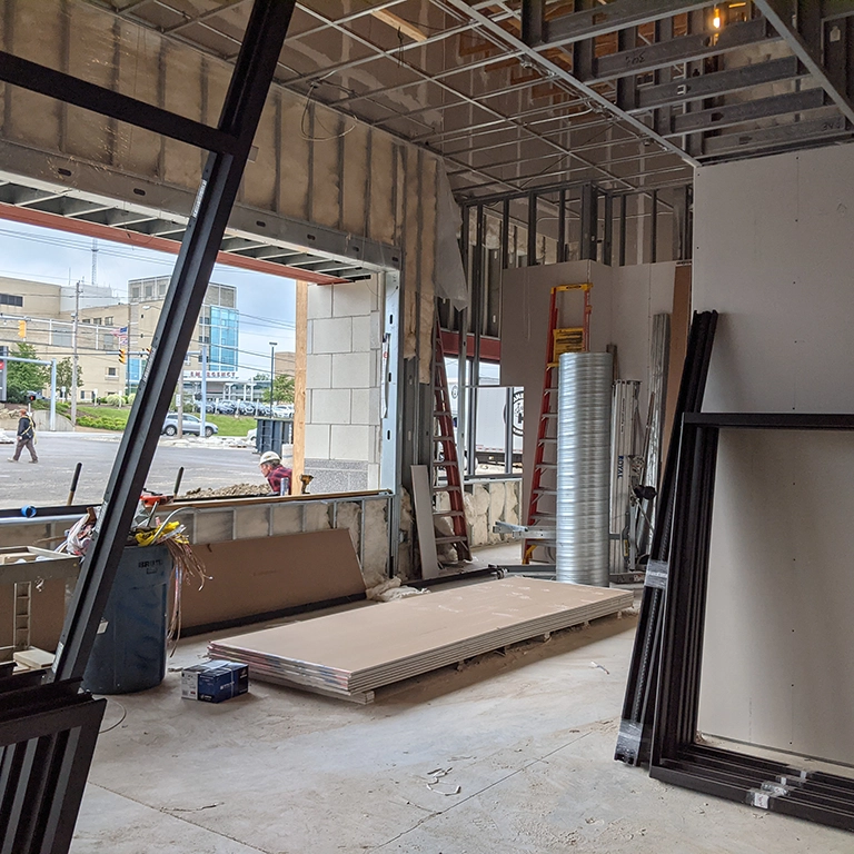 Construction site interior with exposed framing, drywall sheets on the floor, ladders, and building materials scattered around; large open window reveals street and buildings outside.