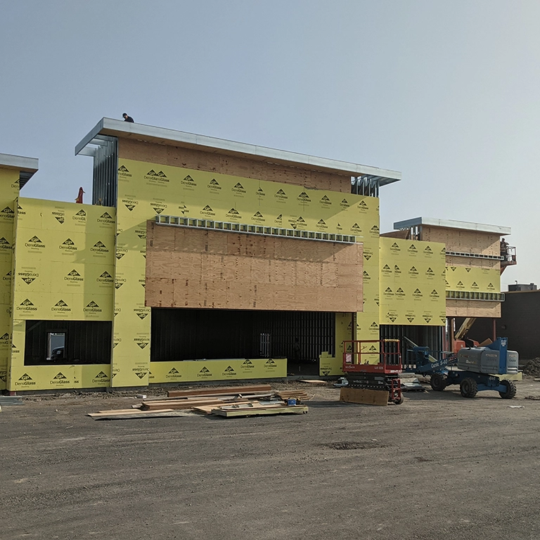 A commercial building under construction with yellow sheathing, boarded windows, construction equipment, and materials on the ground.