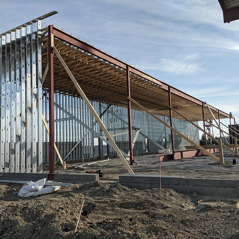 A metal-framed building under construction with exposed beams and wall studs is set on a cleared dirt lot under a blue sky.