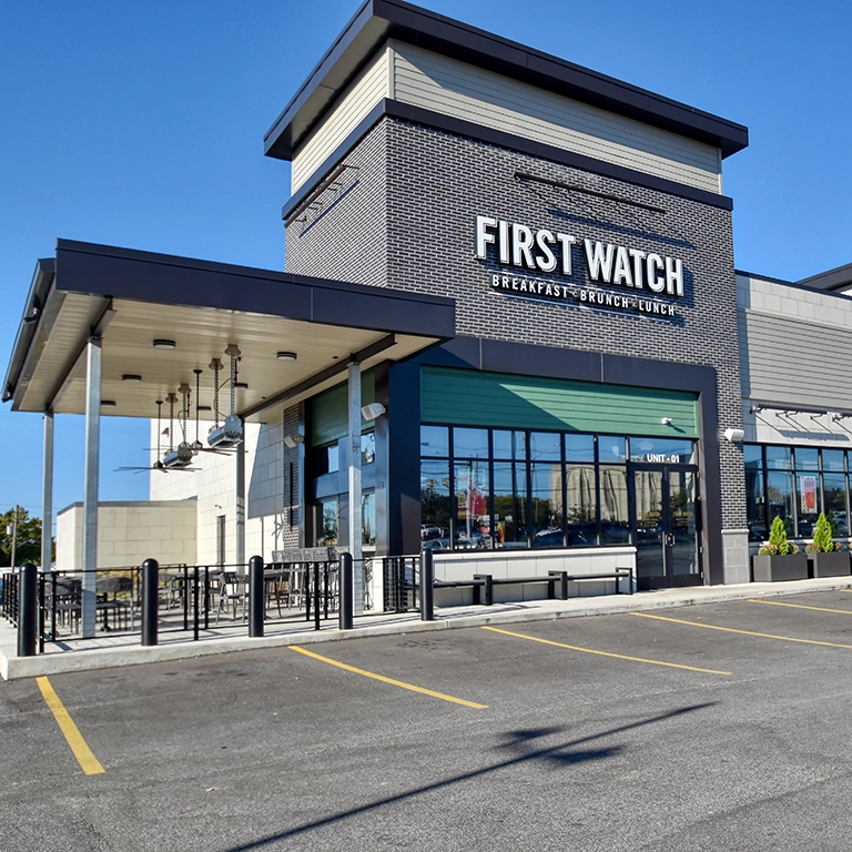 Exterior view of a First Watch restaurant with outdoor seating, large windows, and an adjacent parking lot under a clear sky.