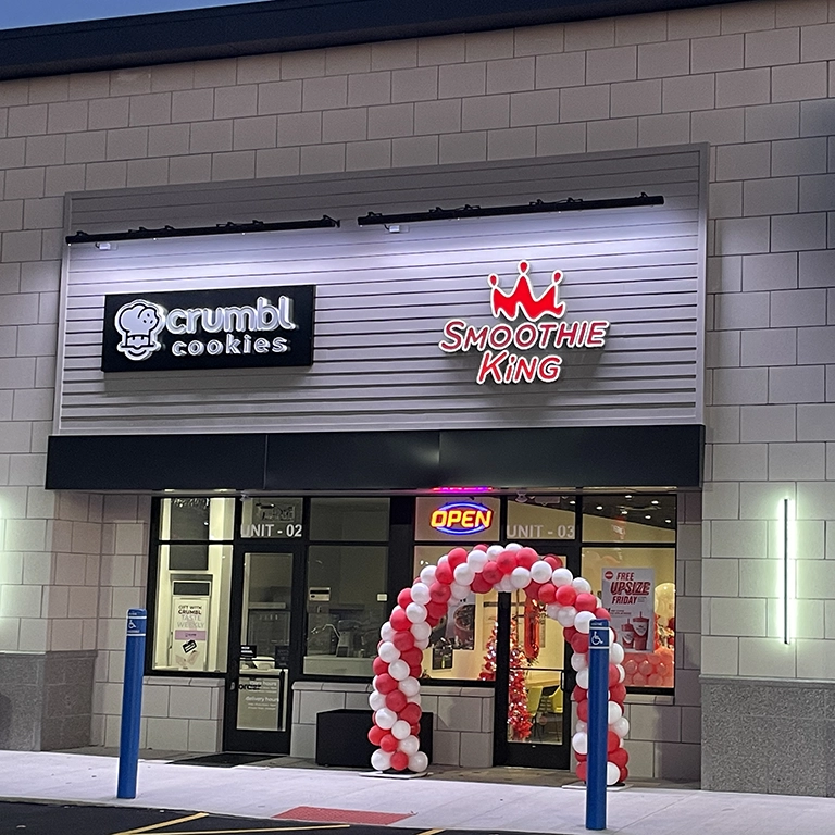Storefront with Crumbl Cookies and Smoothie King signs. A red and white balloon arch stands at the entrance, and an “OPEN” sign is lit above the door.
