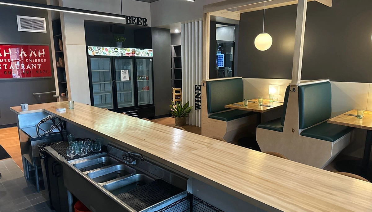 Modern restaurant interior with a long counter, glassware, private booth seating, and a refrigerator in the back.
