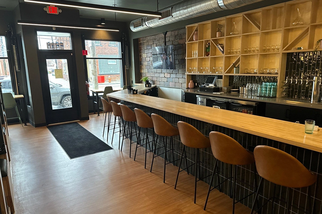 A modern bar with a long wooden counter, brown bar stools, shelves of glasses, and an industrial décor. Large windows and a glass door let in natural light.
