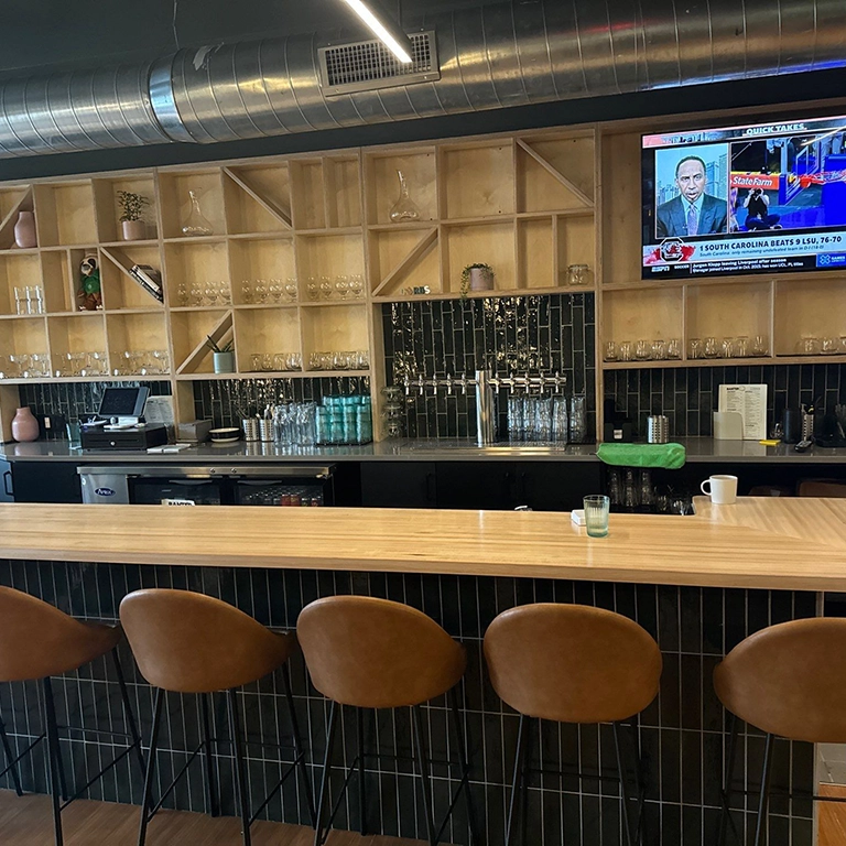 Wooden bar with five brown stools, shelves holding glasses, a TV showing news, and a countertop with a coffee cup, glass, and a green bowl. Modern industrial decor.