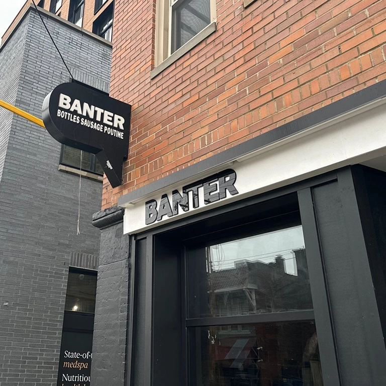 Street view of a brick building with a black sign reading “BANTER BOTTLES SAUSAGE POUTINE” and “BANTER” in black letters above a dark doorway.
