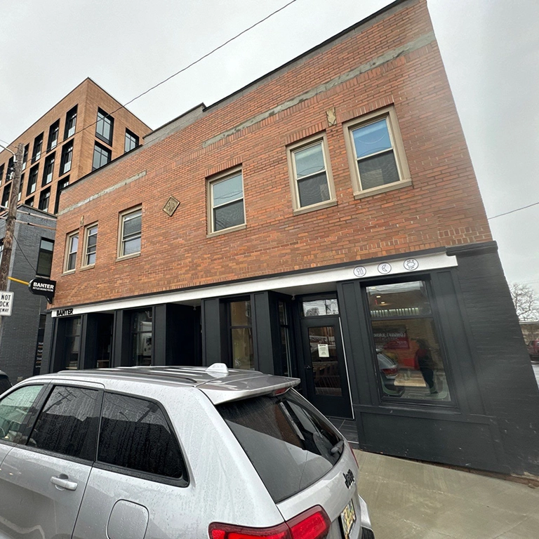 A two-story brick building with large black-framed windows on the ground floor and a silver SUV parked in front on a cloudy day.