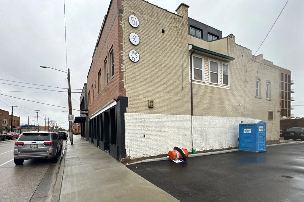 A brick building with restaurant signs, a partially painted wall, a portable toilet, and an overturned orange sign on a wet street.