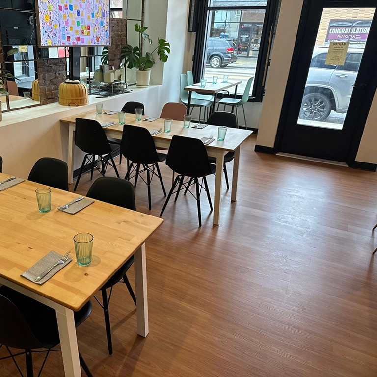 Modern cafe interior with wooden tables, black chairs, place settings, large windows, potted plants, and a view of parked cars outside.