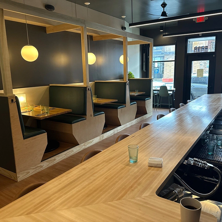 Modern restaurant interior with wooden booths, a long bar, pendant lighting, and glassware on tables. Large front windows provide natural light. No people are present.