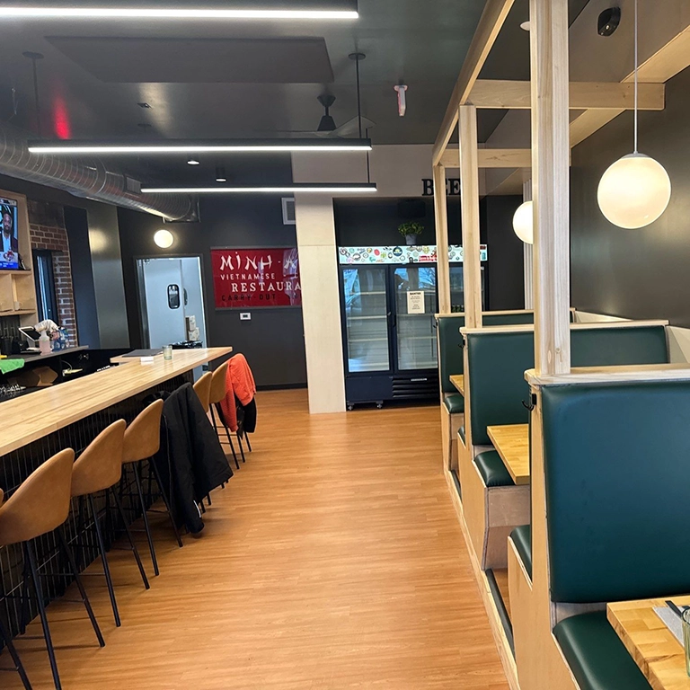 A restaurant interior with wooden floors, green booth seating, bar stools at a counter, and a red sign that says “restaurant” in the background.